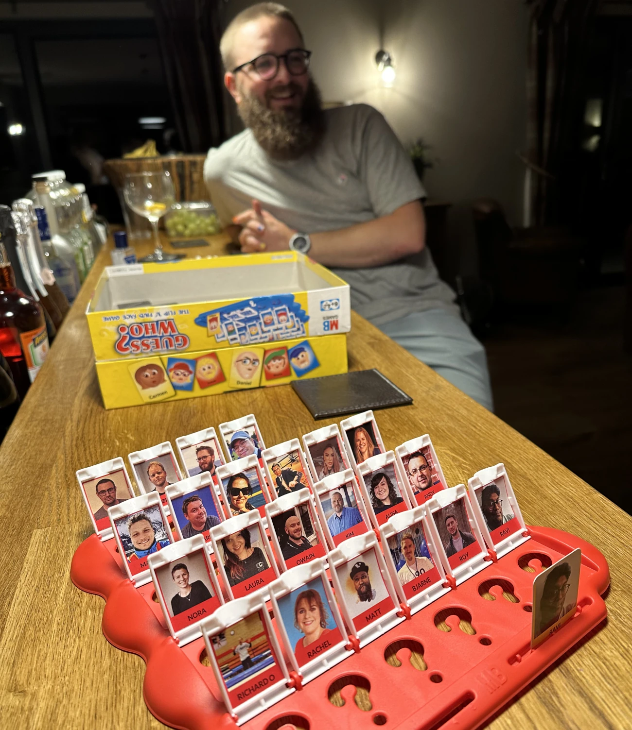 A photograph taken by Joe's Guess Who opponent, with a custom Guess Who board made up of CODECABIN attendees. Joe is sat opposite the photographer at the bar, laughing. Bottles of drink can be seen stacked along the bar.
