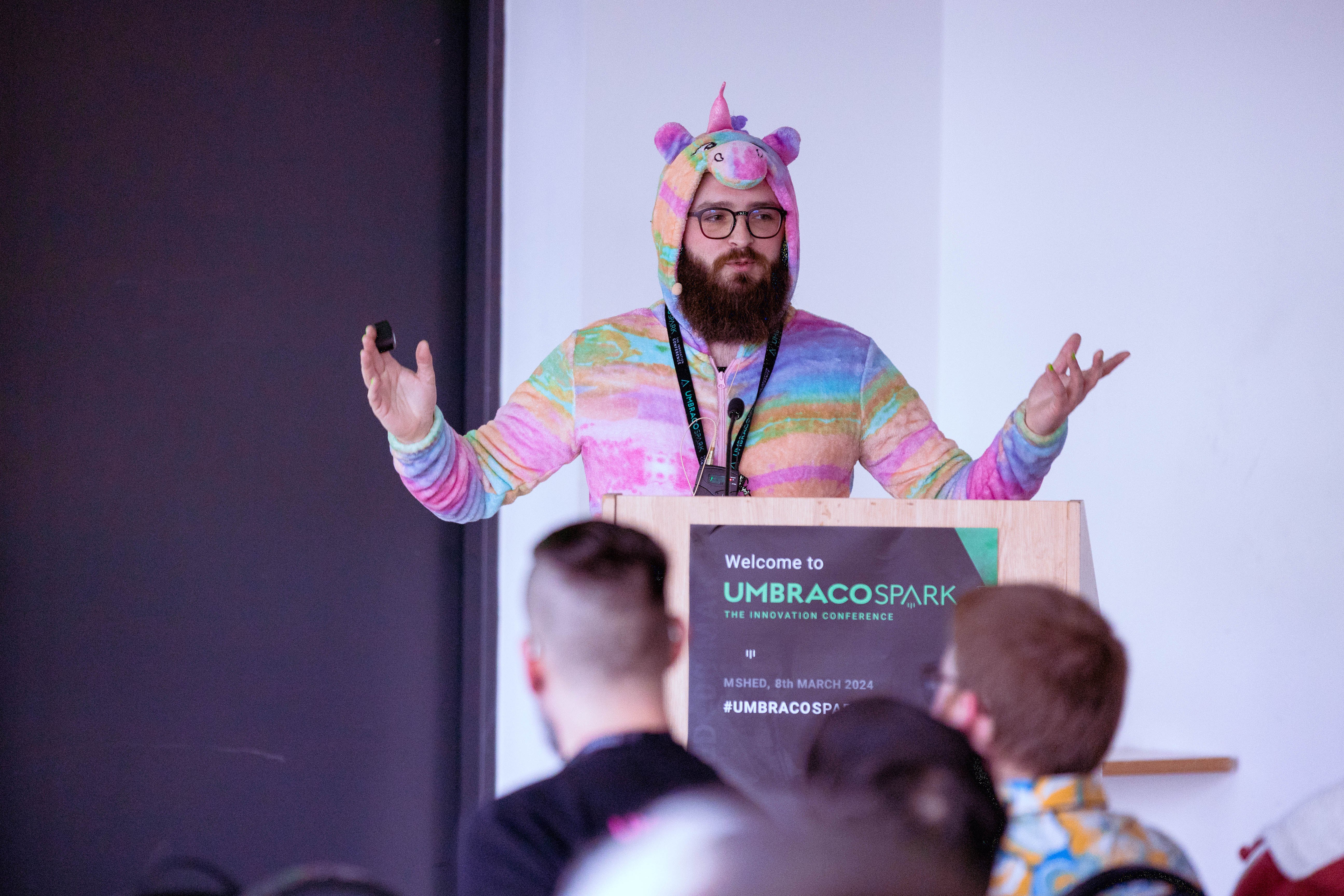 Joe speaking at Spark 2024 wearing a unicorn onesie and stood behind a poduim with the Umbraco Spark logo on it.