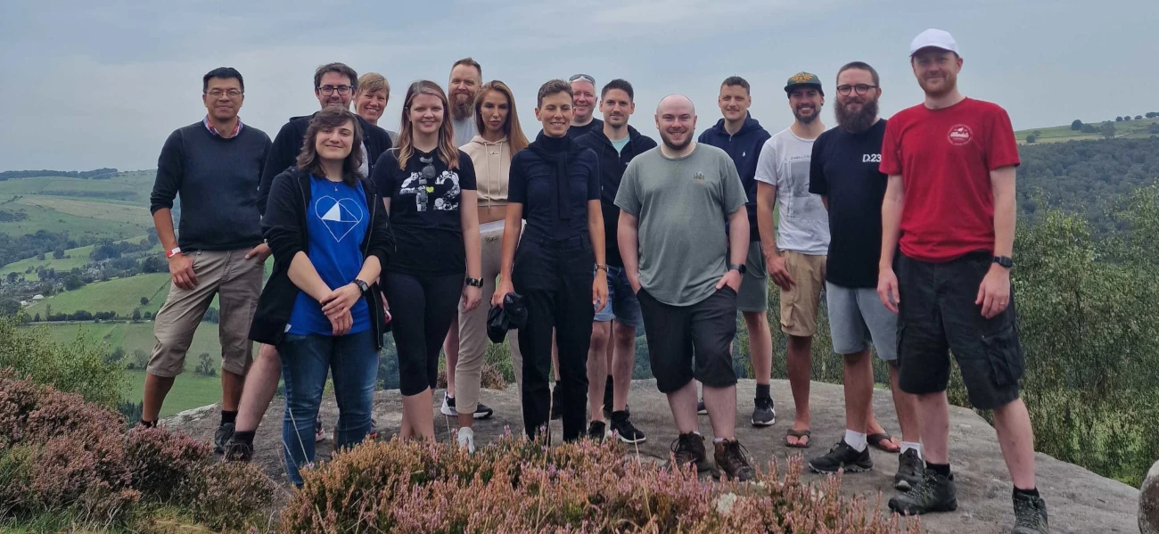 A group photo of those who went on the walk. We're stood on a large flat boulder at the top of Froggatt Edge, with a view of the patchwork of green fields in the valley below.